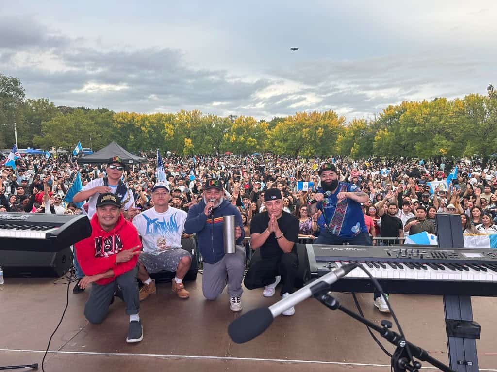 Group of musicians posing on stage with a large cheering crowd at an outdoor concert, keyboard and microphone in front, trees in the background, drone visible in the sky.