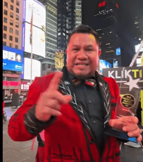 Smiling man in a red suit points at the camera in a busy neon city square, holding an award plaque with a star design.