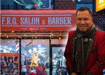 frank lopez migrante emprendedor barber barbero guatemalteco en nueva york originario de cunen quiche 2 – SoyMigrante.com REVISTA Smiling man in a red jacket standing outside a barber shop; a colorful bird flies across the storefront and a scissors-and-comb graphic hovers nearby.