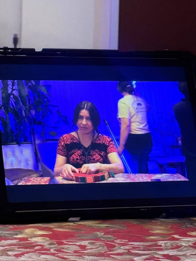 Woman with dark hair in a red patterned top sits at a table, hands on a colorful round woven basket under blue lighting.