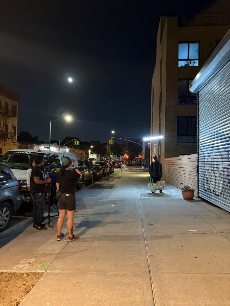 Nighttime city sidewalk with a small group of people filming with a tripod and camera; a man carries shopping bags past a brick building with blue-lit windows.