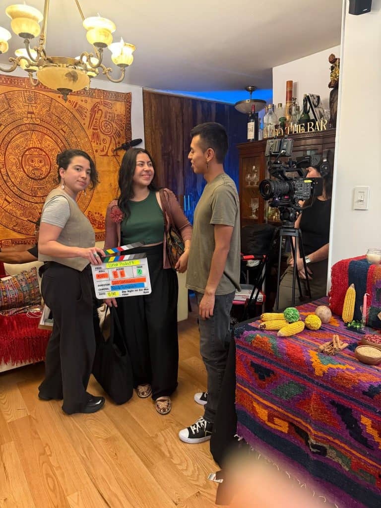 Three people chat in a cozy room; a woman on the left holds a film clapper while a camera on a tripod records them, with a colorful tapestry and decor in the background.