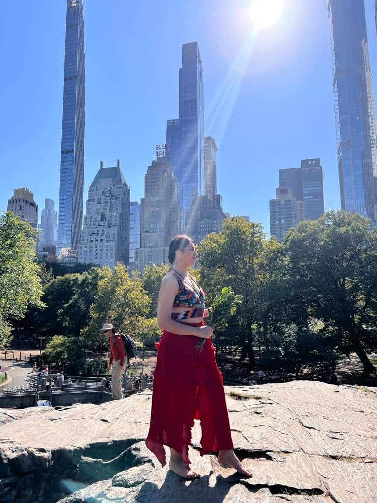 Woman in a colorful sleeveless top and bright red pants stands barefoot on a rocky ledge in a city park, with tall skyscrapers in the background and sunlight streaming down.