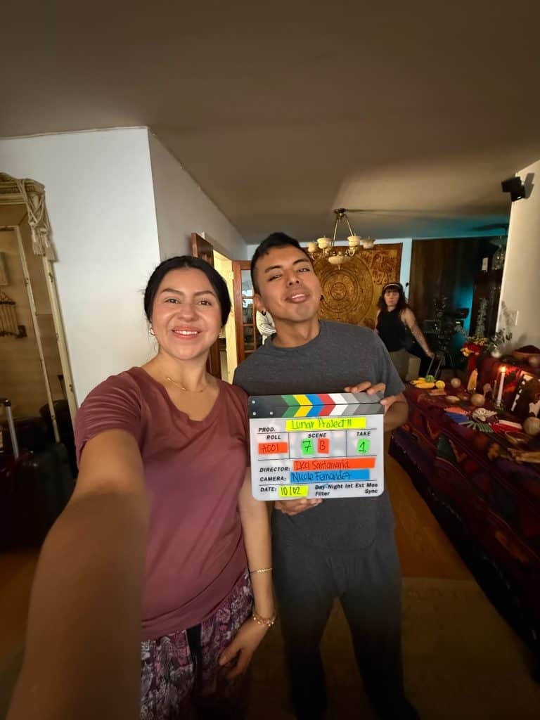 Two people pose for a selfie indoors; the man holds a colorful film clapperboard labeled 'Lunar Project' as a woman stands in the background near a decorated table.