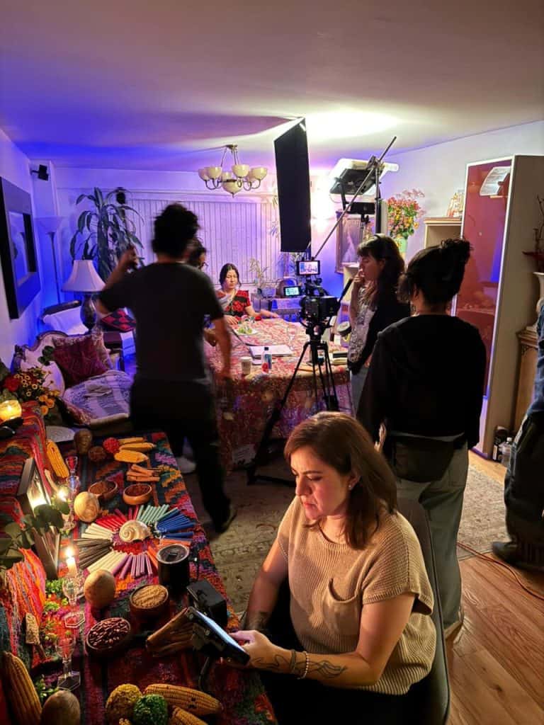 Woman sits at a colorful fabric-covered table with crafts and foods, while a film crew sets up lights and camera in a living-room studio behind her.
