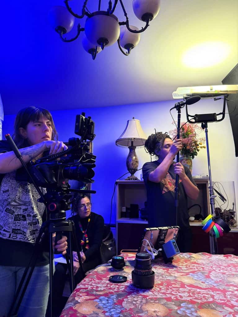 Two people work in a home recording studio with a Sony camera on a tripod, bright blue and yellow lighting, and various lenses and gear on a floral table.