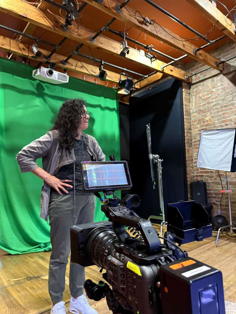 Person with curly hair standing beside a large video camera on a studio set with a green screen behind her.