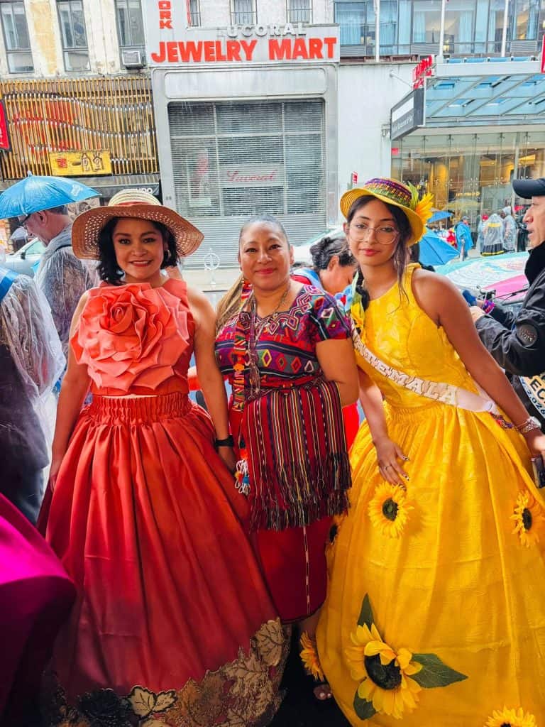 Three women in bright, formal dresses posing on a city street at a parade or festival. Red dress with large flower, patterned dress, and yellow sunflower-accented dress with hats nearby.