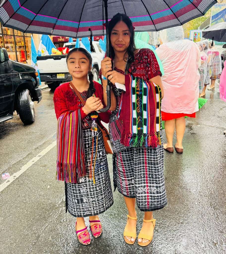 Two women in matching plaid skirts and colorful woven shawls stand in the rain, sharing a large black umbrella on a city street.