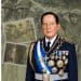 Portrait of a high-ranking military officer in dress uniform with medals and a blue-and-white sash, set against a montage of archival photos behind him.