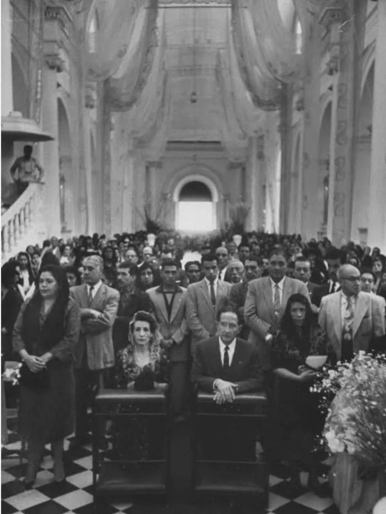 Black-and-white photo of a ceremonial gathering in a church; front row features a man in a suit and a woman seated, with a large crowd standing behind them and arches overhead.