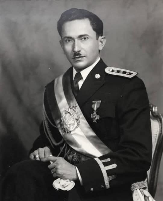 Formal studio portrait of a man in a ceremonial military uniform with a sash, medals, and epaulettes, seated with hands folded.