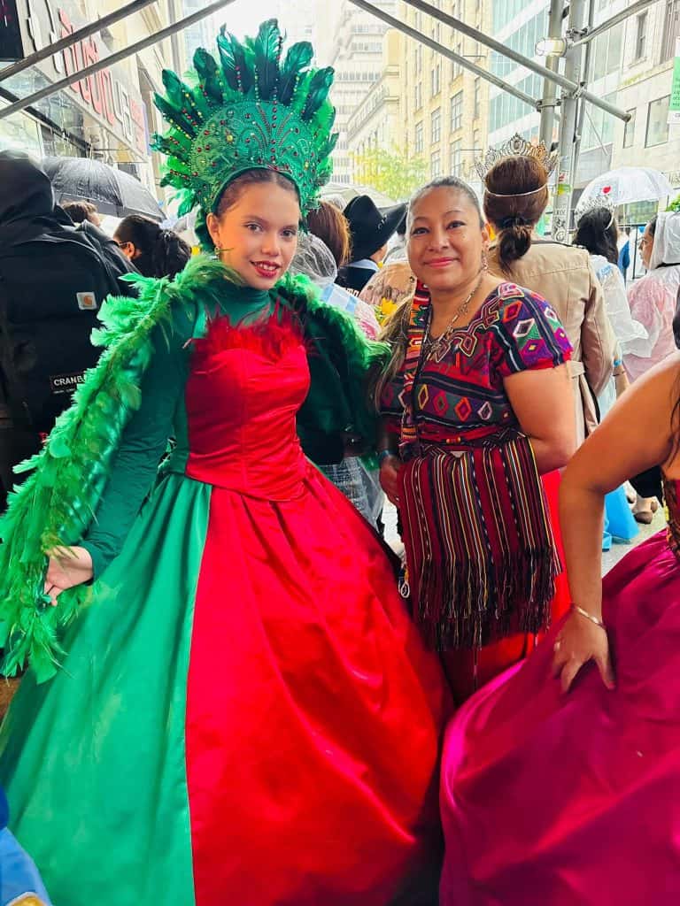 Two women pose for the camera in bright carnival costumes on a crowded city street festival.
