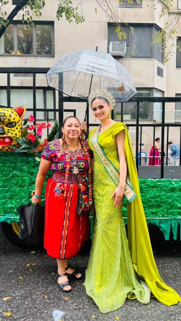 Two women pose at a street parade: a beauty contestant in a lime-green gown and crown, beside a woman in a bright, patterned traditional outfit holding a clear umbrella behind them.