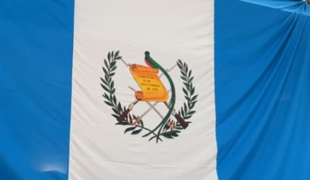 Bandera de Guatemala con el Escudo Nacional, durante una reciente manifestación de migrantes en contra de actos antidemocráticos del Ministerio Público y ciertos jueces. El escudo tiene todas las características reglamentarias. – SoyMigrante.com – SoyMigrante.com