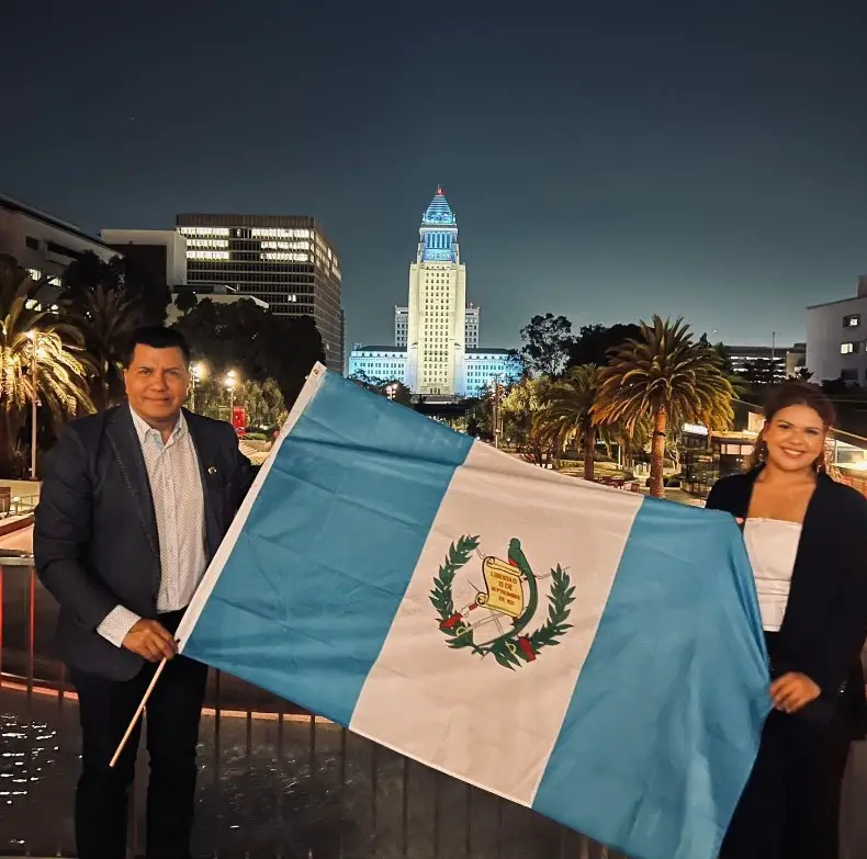 Migrantes chapines posan con la bandera nacional y al fondo, el City Hall de Los Angeles iluminado de azul y blanco, con motivo de la Semana del Guatemalteco – SoyMigrante.com – SoyMigrante.com